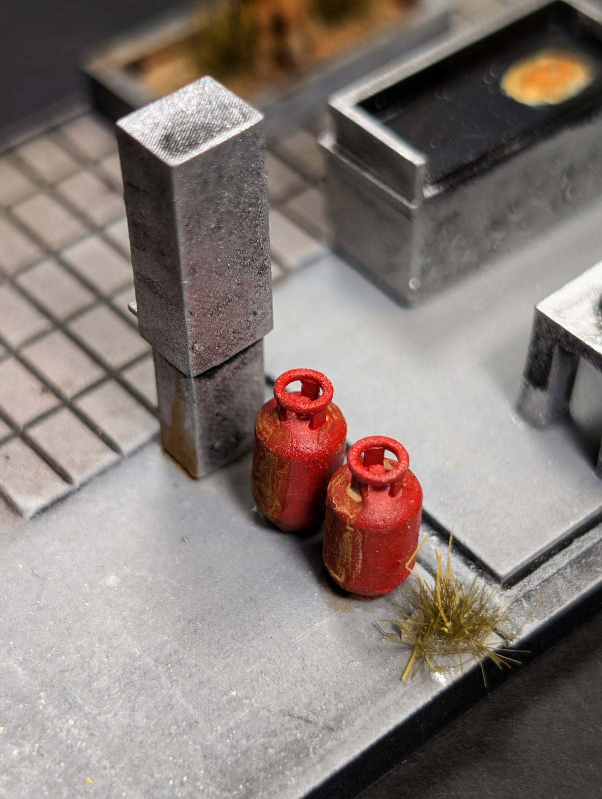 Two miniature red gas bottles with gold lettering, placed on a gray tabletop terrain base alongside a gray metal post and other scale models.