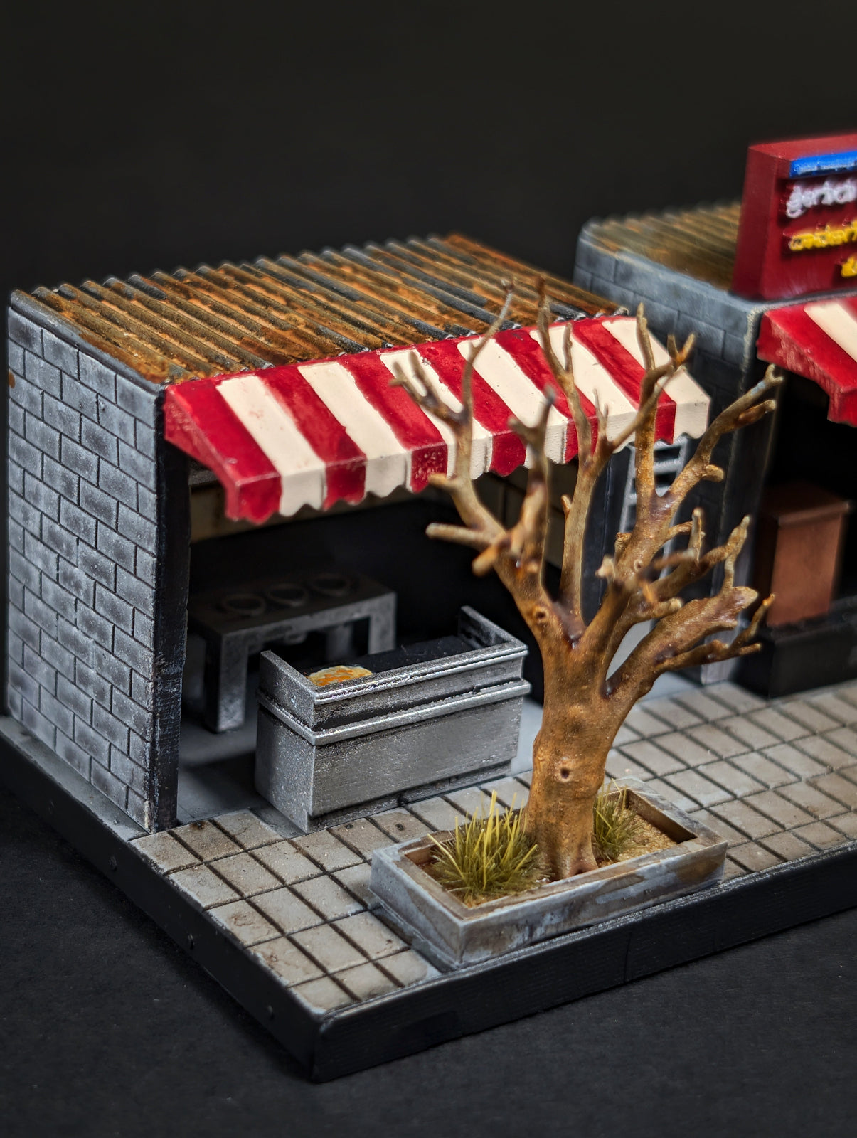 A miniature diorama model of a street market stall with a gray brick facade, a red and white striped awning, tiled floor, and a small bare tree in a planter.