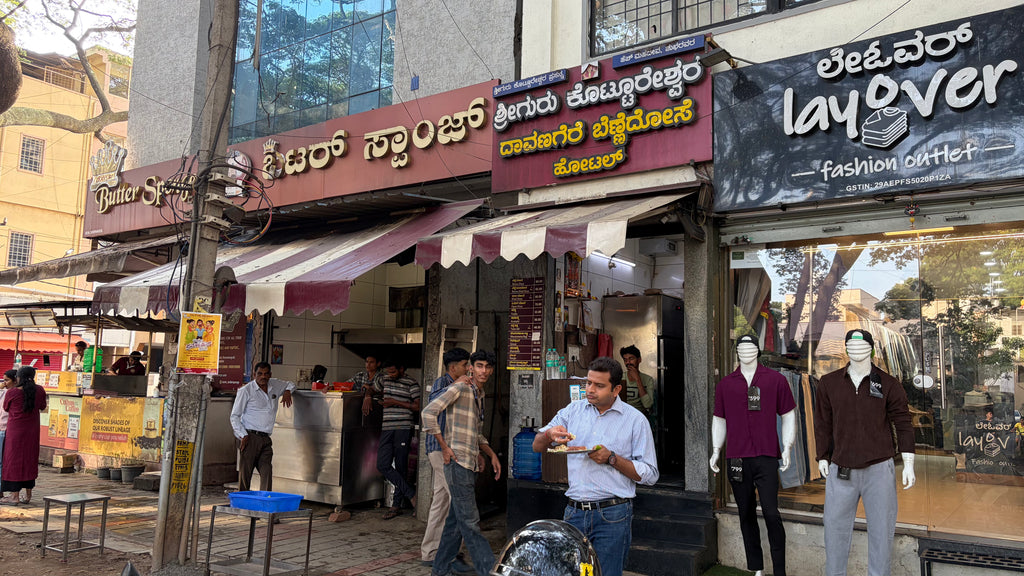 Street scene in front of a retail shop with multiple storefronts, awnings, pedestrians, mannequins, and signage including 'layover fashion outlet'.