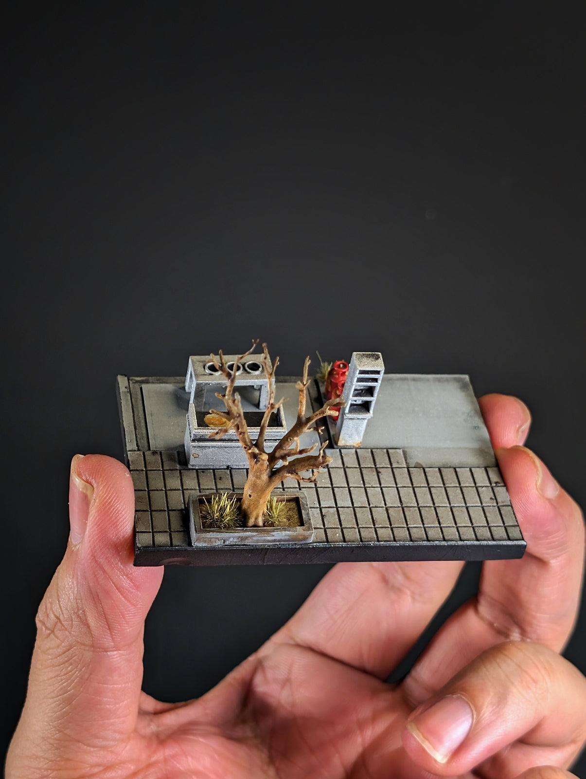 A miniature tabletop terrain piece depicting a barren tree in a planter next to a small structure, mounted on a grid-patterned base and held in two hands against a dark background.