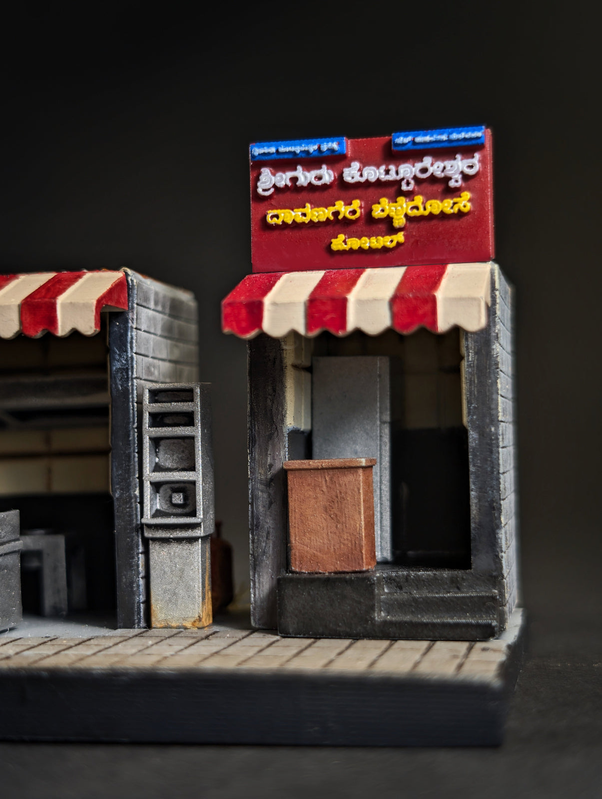 A miniature model of a street shop with a red and cream striped awning, a red signboard with yellow and white text, and a gray brick facade, displayed on a dark background.